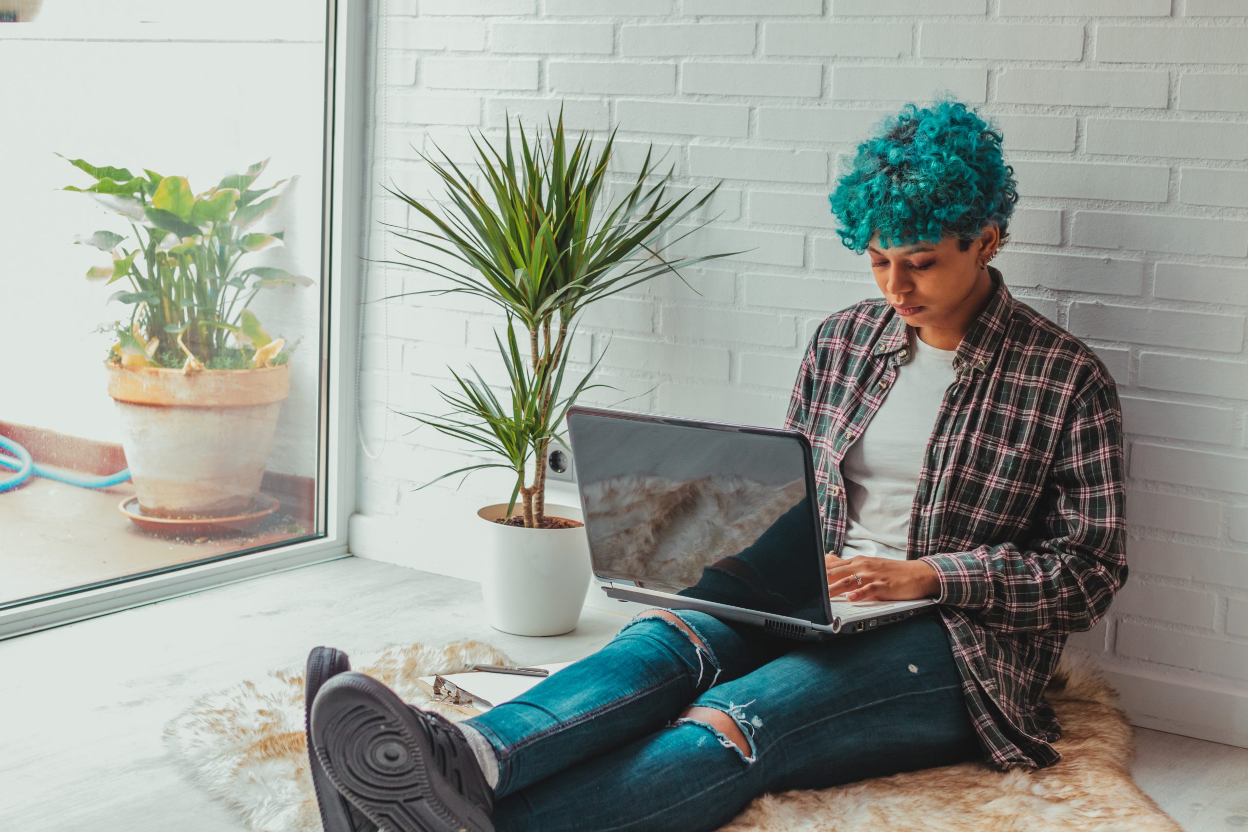 Young person sitting on the floor against a brick wall working on a laptop