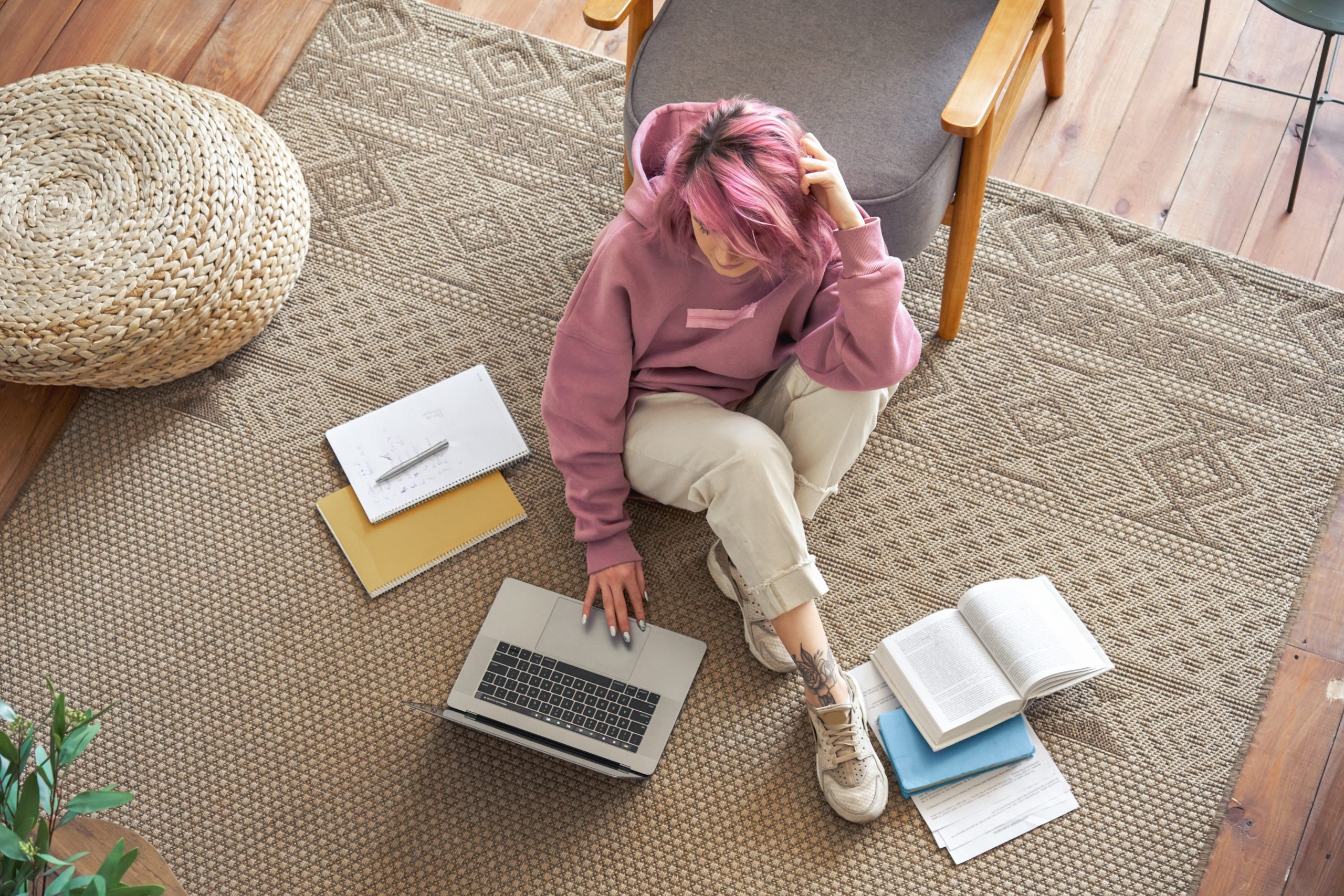 Teenage girl with pink jumper and pink hair sitting on living floor and doing school work on laptop