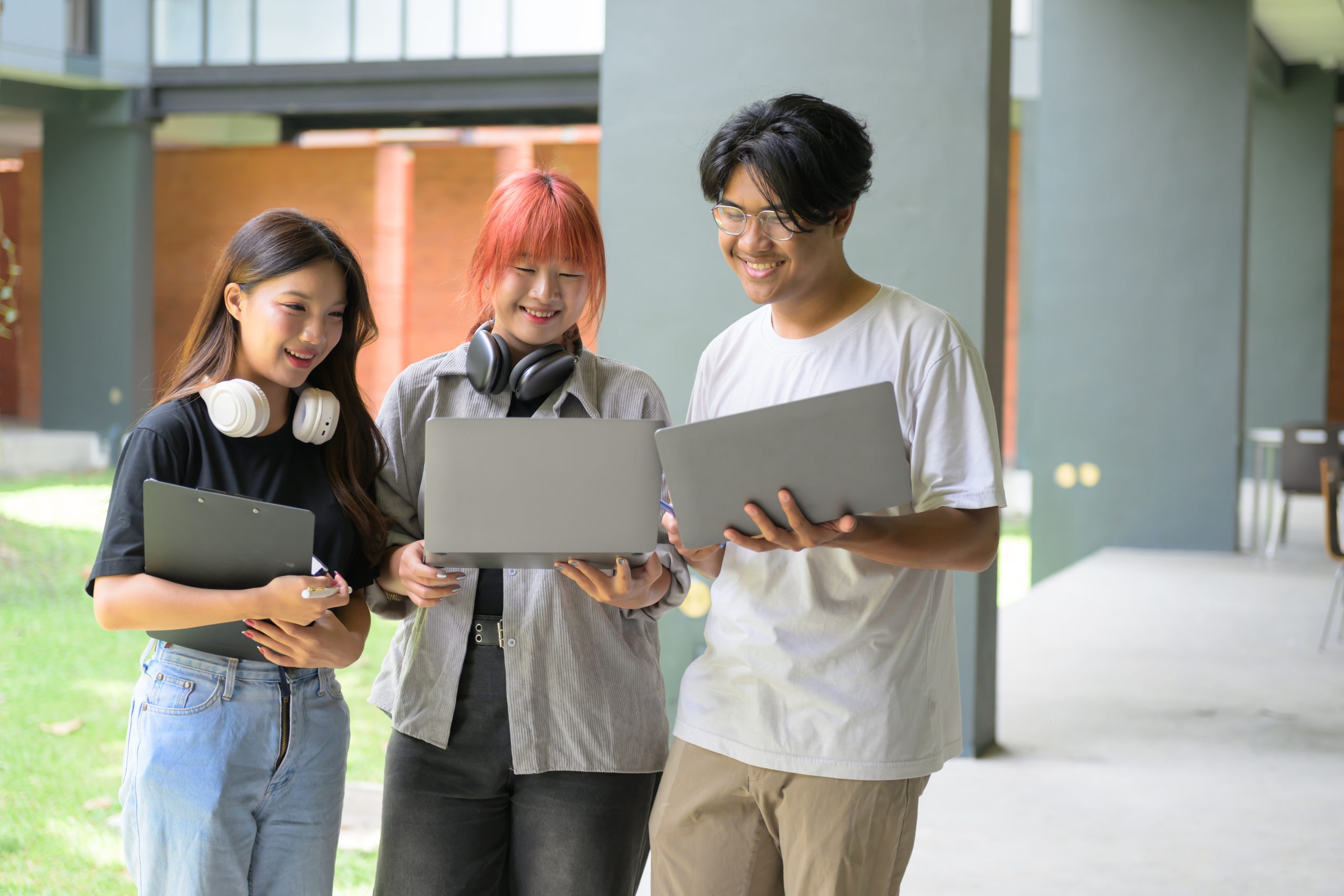 Three students working together with laptops and tablets outside a contemporary university building, showcasing teamwork and technology in education.